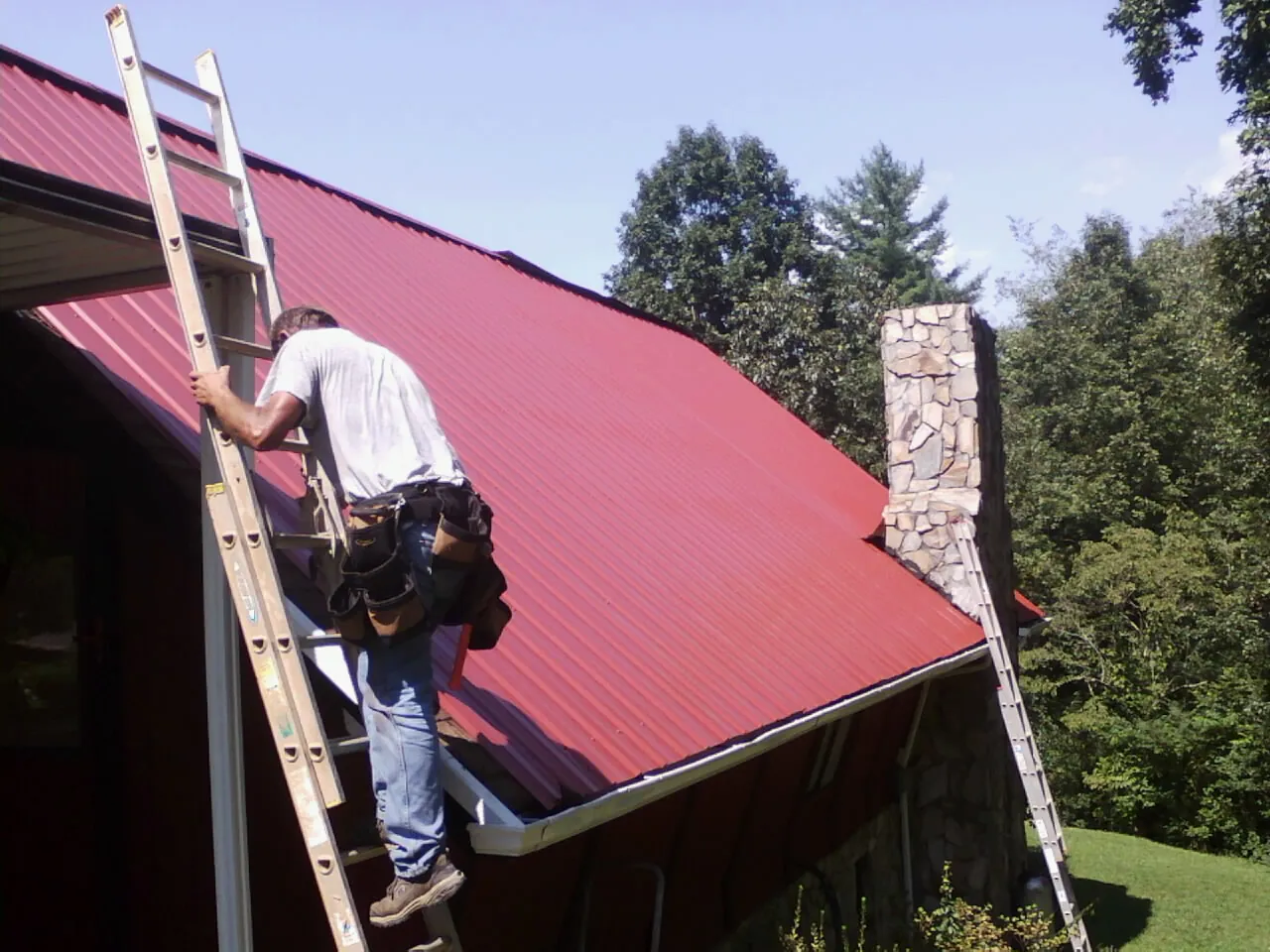 Roofer on a ladder beside a red metal roof and stone chimney — Soffit & Fascia Repair by Roofing Brothers in Edgeley